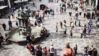 A bustling city square with many people walking and gathering. The ground has a geometric stone pattern, and there is a fountain on the left. Surrounding the square are historic buildings with various architectural styles.