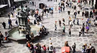 A bustling city square with many people walking and gathering. The ground has a geometric stone pattern, and there is a fountain on the left. Surrounding the square are historic buildings with various architectural styles.