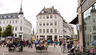 A bustling European city square on a cloudy day, featuring cobblestone streets, people walking, and some with strollers and bicycles. The area is surrounded by historic buildings with a café visible on the right.