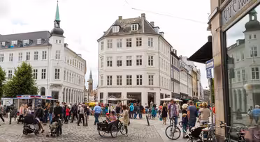 A bustling European city square on a cloudy day, featuring cobblestone streets, people walking, and some with strollers and bicycles. The area is surrounded by historic buildings with a café visible on the right.