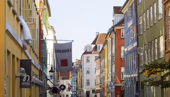 A street in Copenhagen with colorful buildings lining both sides. People are walking and cycling, and cars are parked along the street. A flag with København K hangs across the street. The sky is clear and blue.