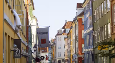 A street in Copenhagen with colorful buildings lining both sides. People are walking and cycling, and cars are parked along the street. A flag with København K hangs across the street. The sky is clear and blue.
