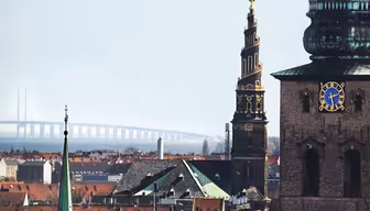 Skyline view of Copenhagen featuring the Church of Our Saviour with its spiral tower and ornate spire. Nearby is a historic brick building with a clock. In the background, the Øresund Bridge stretches across the water under a clear sky.