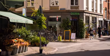 A sunny street corner. On the left, a flower shop with plants and bouquets. Bicycles are parked near a shop called DORA. People are strolling nearby, and an ice cream cone is depicted on a chalkboard. The buildings are decorated with greenery.