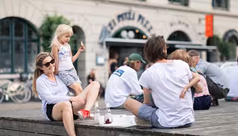 A group of people sitting on a wooden platform outside, with a woman wearing sunglasses and a child with blonde hair. In the background, theres a building with the text pizza visible. Bottles and food are on the ground.