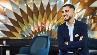 A man in a suit smiles while sitting in a modern, stylish lounge with geometric wall art. He has his arms crossed and wears a name badge. The setting includes blue chairs and a reflective, artistic background.