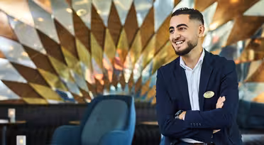 A man in a suit smiles while sitting in a modern, stylish lounge with geometric wall art. He has his arms crossed and wears a name badge. The setting includes blue chairs and a reflective, artistic background.
