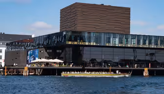 A modern, glass-fronted building with a unique textured upper section sits by the water. A long, open-top boat with tourists passes in front of it. Clear skies and scattered clouds are visible in the background.
