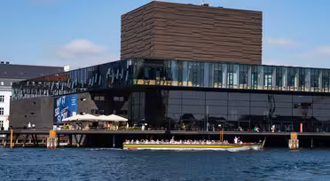 A modern, glass-fronted building with a unique textured upper section sits by the water. A long, open-top boat with tourists passes in front of it. Clear skies and scattered clouds are visible in the background.