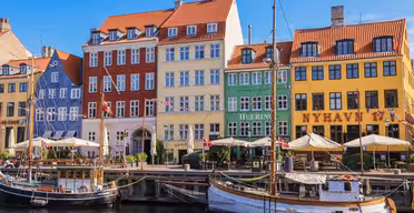 Colorful historic buildings line the Nyhavn waterfront in Copenhagen, Denmark. The structures are painted in red, yellow, and green, with outdoor cafes and sailing boats docked along the canal under a clear blue sky.