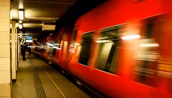 A red train speeds through a dimly lit underground station. The motion creates a blur effect, emphasizing the trains speed. The platform is mostly empty, with a few distant figures visible under fluorescent lights.
