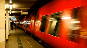 A red train speeds through a dimly lit underground station. The motion creates a blur effect, emphasizing the trains speed. The platform is mostly empty, with a few distant figures visible under fluorescent lights.