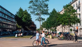 A bustling city scene with people walking and sitting in a park. Two women stroll past with a bicycle. Trees line the pathway, and contemporary buildings flank both sides of the street. The sky is clear and sunny.