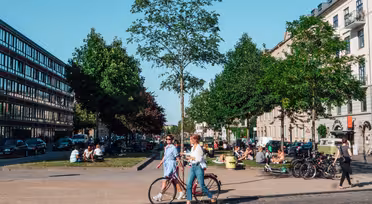 A bustling city scene with people walking and sitting in a park. Two women stroll past with a bicycle. Trees line the pathway, and contemporary buildings flank both sides of the street. The sky is clear and sunny.