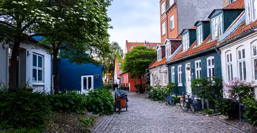 A charming cobblestone street lined with colorful, historic townhouses. Trees and bushes with flowers add greenery, and bicycles are parked along the road. The sky is overcast, providing a calm, picturesque atmosphere.