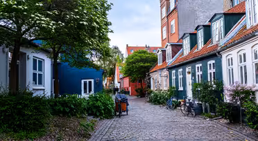 A charming cobblestone street lined with colorful, historic townhouses. Trees and bushes with flowers add greenery, and bicycles are parked along the road. The sky is overcast, providing a calm, picturesque atmosphere.