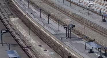 An aerial view of an empty train station with multiple parallel tracks and platforms. There are small shelters and signage on the platforms, with no people present. The setting appears bright and clear.