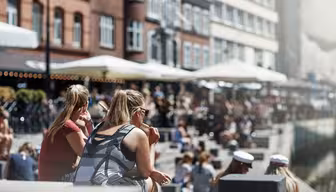 People sit along a canal on a sunny day with brick buildings and outdoor cafes along the water. The scene is lively, with people enjoying the weather, some wearing sunglasses and others in white caps.