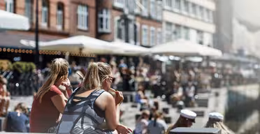 People sit along a canal on a sunny day with brick buildings and outdoor cafes along the water. The scene is lively, with people enjoying the weather, some wearing sunglasses and others in white caps.