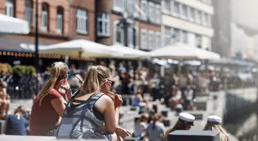 People sit along a canal on a sunny day with brick buildings and outdoor cafes along the water. The scene is lively, with people enjoying the weather, some wearing sunglasses and others in white caps.