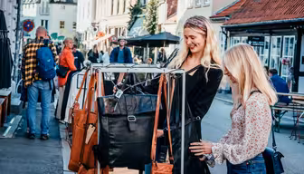 Two women smile while browsing handbags on a street rack in a bustling outdoor market. They are surrounded by people walking and looking at various stalls, with charming buildings lining the street under a clear sky.