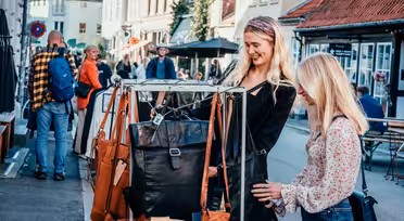Two women smile while browsing handbags on a street rack in a bustling outdoor market. They are surrounded by people walking and looking at various stalls, with charming buildings lining the street under a clear sky.