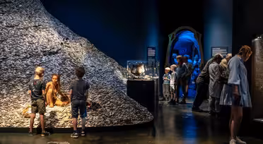 A dimly lit museum exhibit features a mound covered in shells, where a woman sits posing as a display. Nearby, children watch as visitors explore other exhibits, including a glass case and a glowing blue archway leading to another area.