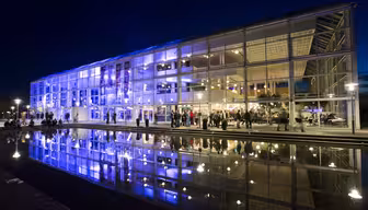 A modern building with glass walls is illuminated in purple and blue lights at night. Reflections are visible in a calm water surface in front. People are gathered outside and inside, appearing to enjoy an event.