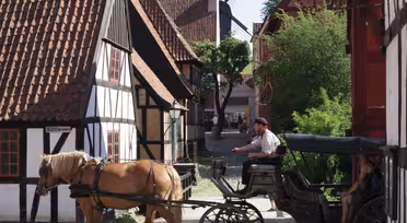 A horse-drawn carriage travels down a cobblestone street lined with traditional half-timbered houses. A windmill is visible in the background, and trees dot the surroundings under a clear blue sky.