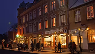 A lively street scene at dusk features people walking past a warmly lit building adorned with festive lights and decorations. The building houses shops with bright displays, and a deep blue evening sky forms the backdrop.