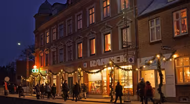 A lively street scene at dusk features people walking past a warmly lit building adorned with festive lights and decorations. The building houses shops with bright displays, and a deep blue evening sky forms the backdrop.