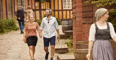 A smiling couple walks hand in hand along a cobblestone street with timber-framed buildings. A person in traditional clothing is in the foreground. Greenery and warm, sunny weather surround the scene.