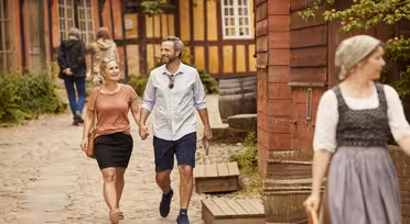 A smiling couple walks hand in hand along a cobblestone street with timber-framed buildings. A person in traditional clothing is in the foreground. Greenery and warm, sunny weather surround the scene.