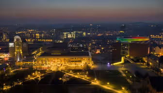 Aerial view of a cityscape at dusk. The skyline is illuminated with colorful lights, highlighting a circular light installation on a building. Soft evening glow casts a tranquil atmosphere over the urban landscape.