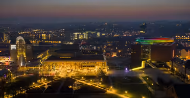 Aerial view of a cityscape at dusk. The skyline is illuminated with colorful lights, highlighting a circular light installation on a building. Soft evening glow casts a tranquil atmosphere over the urban landscape.