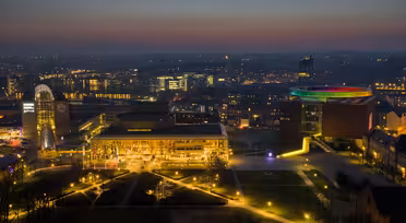 Aerial view of a cityscape at dusk. The skyline is illuminated with colorful lights, highlighting a circular light installation on a building. Soft evening glow casts a tranquil atmosphere over the urban landscape.