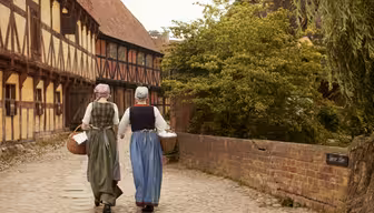 Two women in historical clothing walk on a cobblestone path. One wears a green dress, and the other a blue dress, both with aprons and headscarves. They carry wicker baskets through a medieval-style village, with timber-framed buildings and a brick wall.