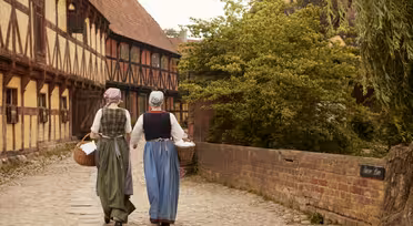 Two women in historical clothing walk on a cobblestone path. One wears a green dress, and the other a blue dress, both with aprons and headscarves. They carry wicker baskets through a medieval-style village, with timber-framed buildings and a brick wall.