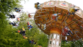 A colorful spinning swing ride at an amusement park, with people enjoying the ride. The swings are suspended from a decorative central structure. The background is filled with green trees, suggesting a lively and vibrant atmosphere.