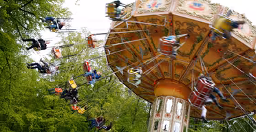 A colorful spinning swing ride at an amusement park, with people enjoying the ride. The swings are suspended from a decorative central structure. The background is filled with green trees, suggesting a lively and vibrant atmosphere.