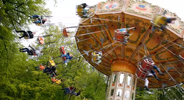 A colorful spinning swing ride at an amusement park, with people enjoying the ride. The swings are suspended from a decorative central structure. The background is filled with green trees, suggesting a lively and vibrant atmosphere.