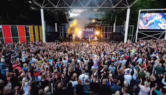 A large outdoor concert with a band performing on stage under a canopy. The audience, consisting of diverse people, stands with hands raised, enjoying the music. Multi-colored panels and a screen flank the stage. Trees are visible in the background.