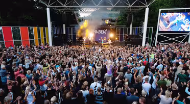 A large outdoor concert with a band performing on stage under a canopy. The audience, consisting of diverse people, stands with hands raised, enjoying the music. Multi-colored panels and a screen flank the stage. Trees are visible in the background.