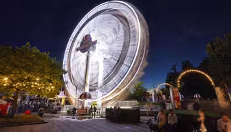 A nighttime scene of an amusement park with a large, illuminated Ferris wheel spinning, creating light trails. The park is bustling with visitors, and decorative lights are strung on trees and arches. People sit and stroll around the area.