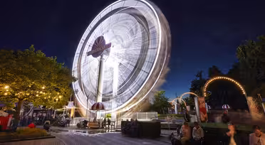 A nighttime scene of an amusement park with a large, illuminated Ferris wheel spinning, creating light trails. The park is bustling with visitors, and decorative lights are strung on trees and arches. People sit and stroll around the area.