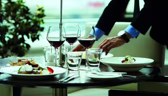 A table set in a restaurant with elegant dishes, including plates of salad and wine glasses. A person in formal attire adjusts the table settings. Background features a plant and soft lighting, creating a sophisticated ambiance.