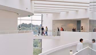 Spiral staircase with people walking along a curved, multi-level walkway inside a modern, white building. Large windows offer views of greenery outside.