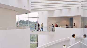 Spiral staircase with people walking along a curved, multi-level walkway inside a modern, white building. Large windows offer views of greenery outside.