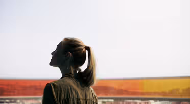 A person with a ponytail stands in silhouette, gazing outward from a window with a tint of red and orange hues. The view outside shows a distant cityscape under a bright sky.