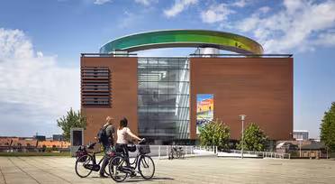 Two people stand with bicycles in front of a modern, red-brick building with large glass windows. A colorful, circular walkway is on the roof. The sky is partly cloudy, and the sun illuminates the scene.